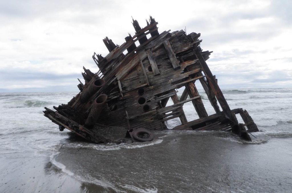 A shipwreck on a sandy beach where only the hull of the wooden ship is left and it's sinking into the wet sand. The sky and ocean in the background are grey.